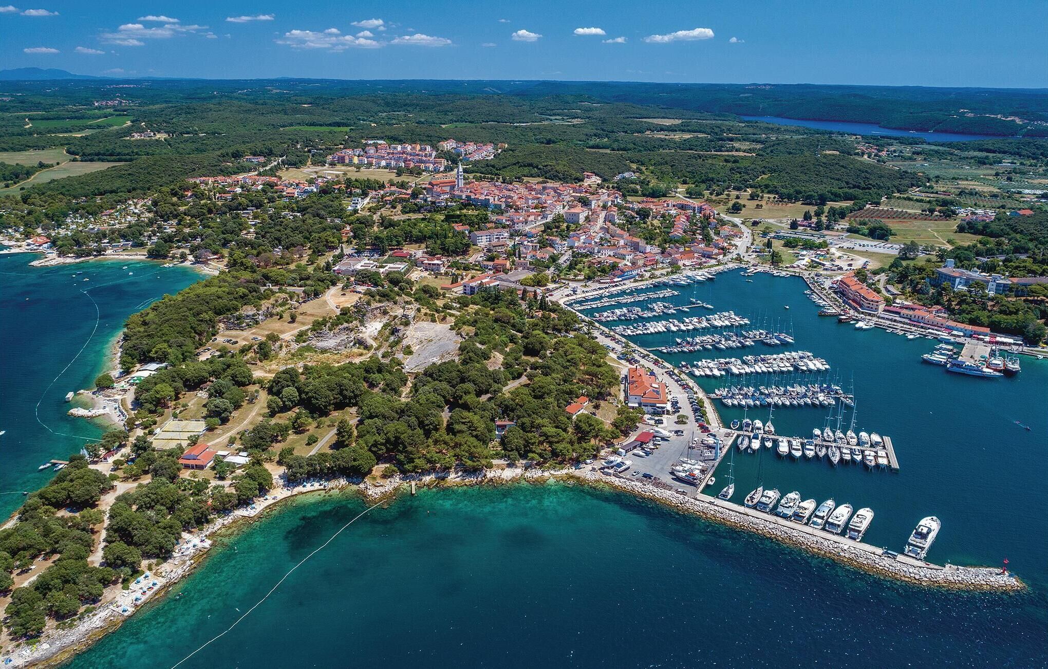 Vista aérea de un pueblo costero con un puerto deportivo, abundante vegetación y edificios residenciales.