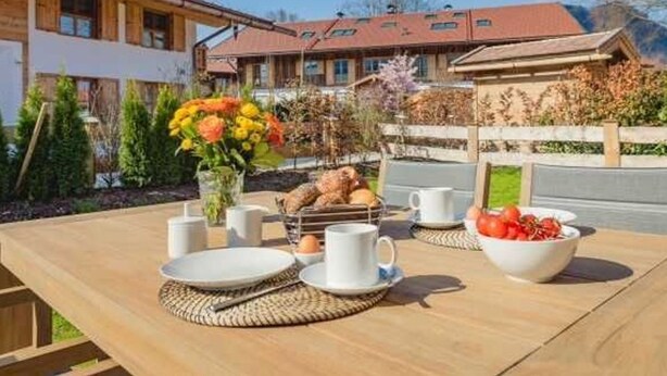 Ein Holztisch gedeckt mit Tellern, Tassen und einer Obstschale, mit Blick auf ein Gebäude und einen Garten im Hintergrund.