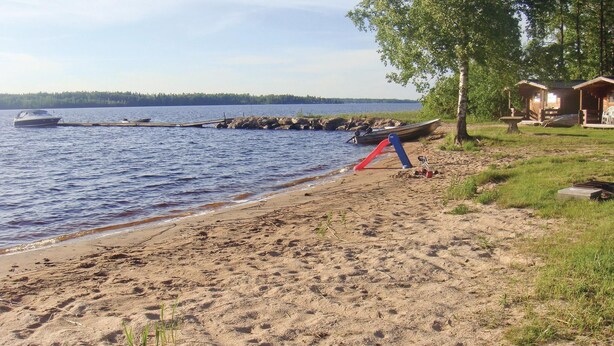 A sandy beach with a boat docked on the water, a small dock, and a row of cabins in the background.