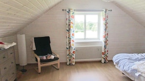 A bedroom with a sloped ceiling, a window with floral curtains, a bed, a chair, a radiator, and a dresser.