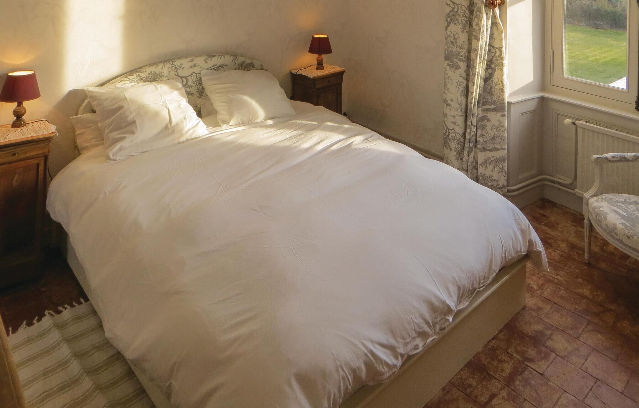 Un lit bien fait, avec du linge de lit blanc, deux lampes de chevet, une chaise et une fenêtre donnant sur un paysage verdoyant.