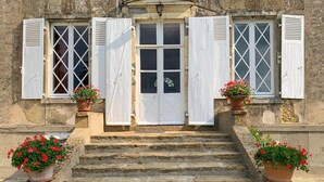 Un bâtiment en pierre, avec un escalier en pierre menant à une porte blanche à volets. De part et d’autre de la porte, on trouve deux plantes en pot ornées de fleurs rouges.