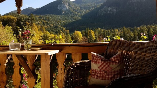 A wooden balcony with wicker furniture, a table with a vase of flowers, and a view of forested mountains. - Apartment Wallberg - Böcklhof, Kreuth-Brunnbichl (Kreuth)
