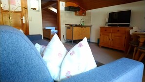 A living room with a sofa, a television, a kitchen area with wooden cabinets, and a radiator. - Apartment Wallberg - Böcklhof, Kreuth-Brunnbichl (Kreuth)