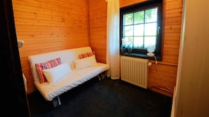 A small room with a wooden interior, a white sofa with striped pillows, a window with a view of greenery, and a radiator. - Cozy Eifelhäuschen in the green near Nürburgring - (Hoffeld)