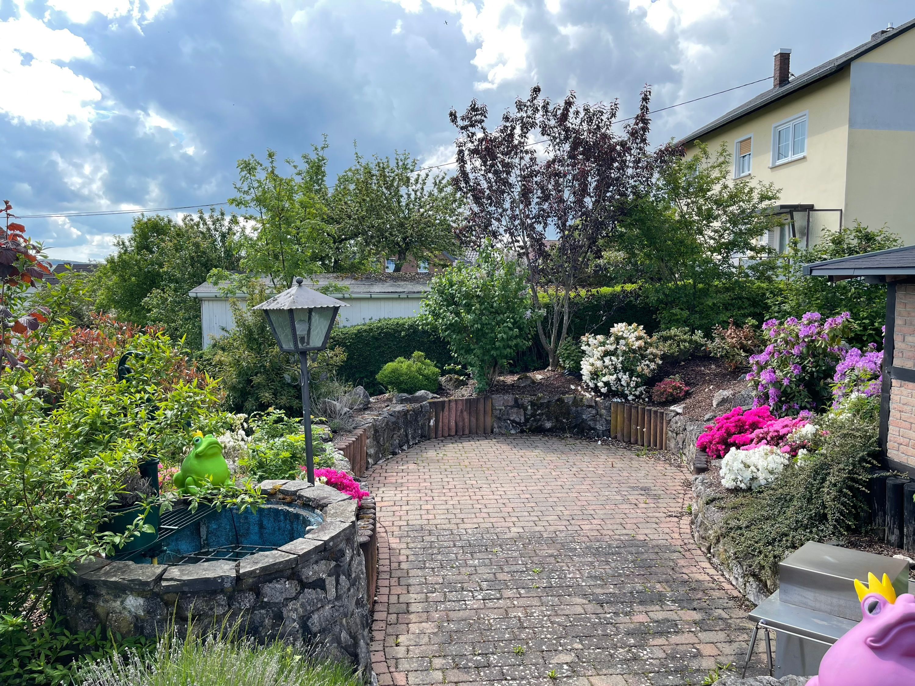 A paved garden path with a stone wall on one side and a variety of plants and flowers on the other.