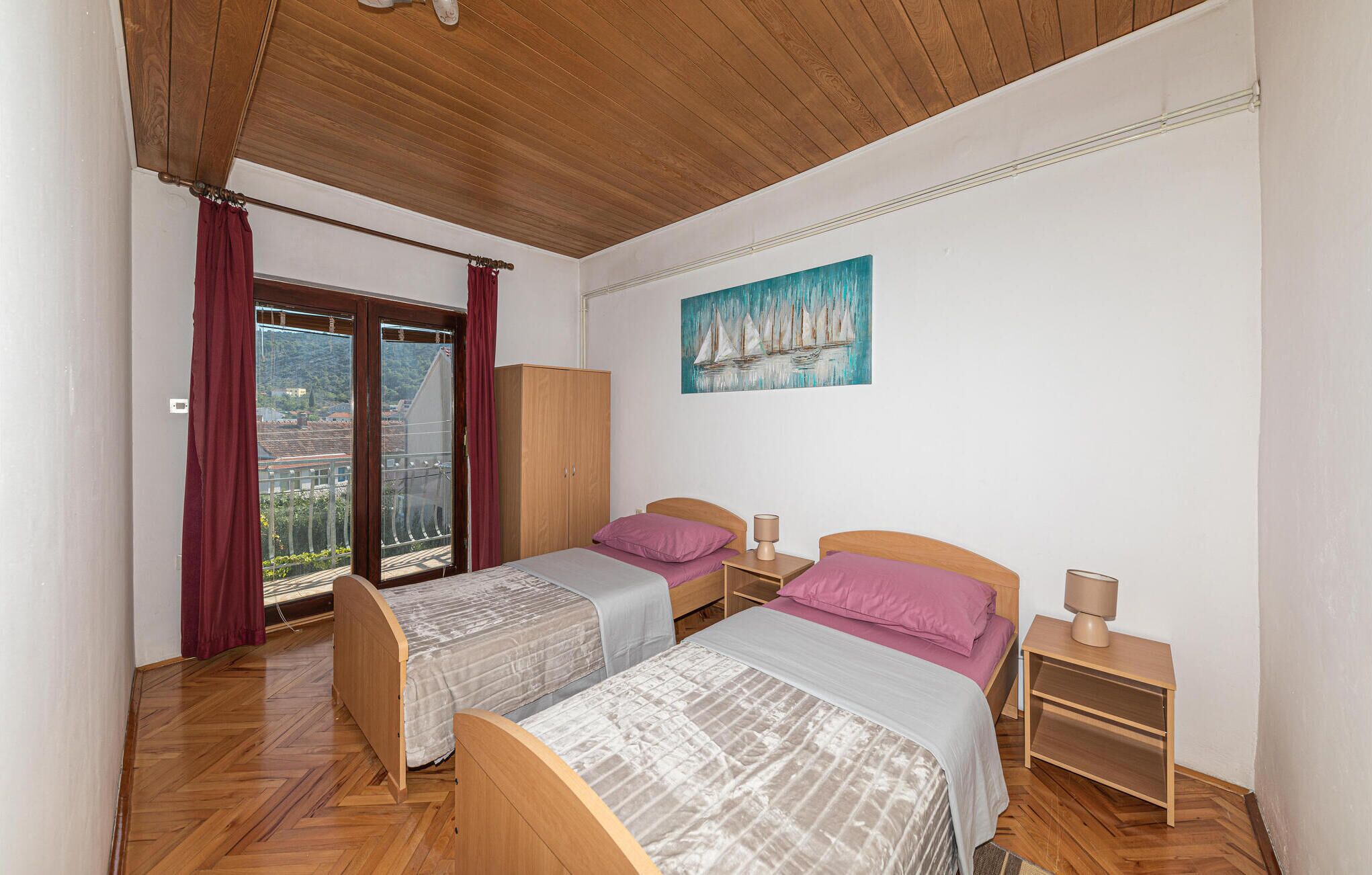 A bedroom with two beds, a wooden wardrobe, a painting of sailboats, and a balcony with red curtains.