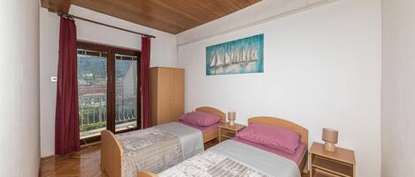 A bedroom with two beds, a wooden wardrobe, a painting of sailboats, and a balcony with red curtains.