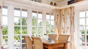 A dining area with a wooden table and wicker chairs, large windows with floral curtains, and a view of greenery outside. - Rasmus 1 from A&M - Rasmus 1 (Kampen (Sylt))