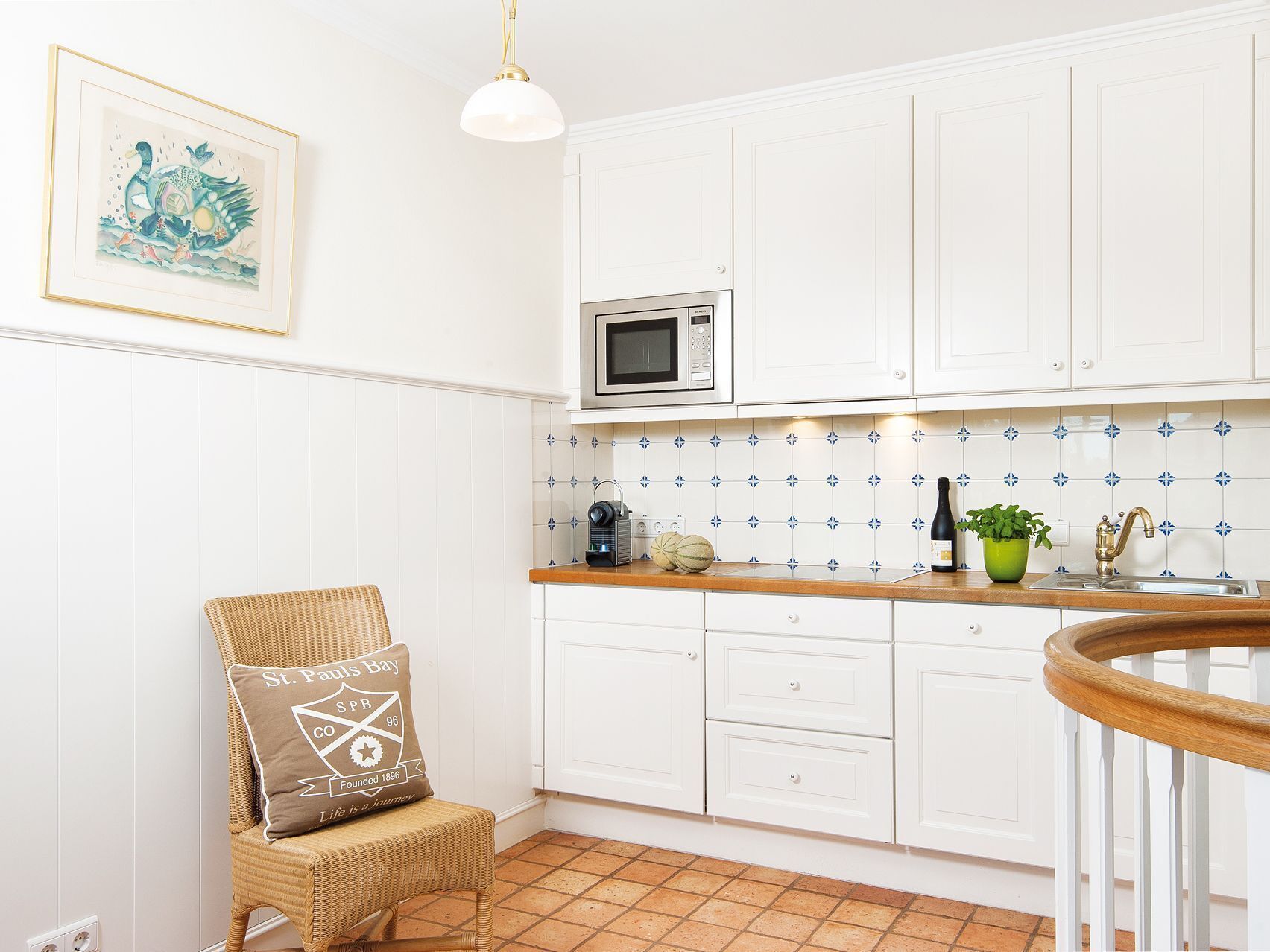 A kitchen with white cabinets, a microwave, a sink, and a wicker chair with a pillow.