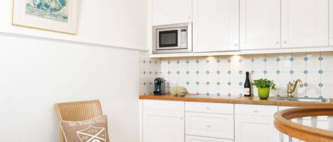 A kitchen with white cabinets, a microwave, a sink, and a wicker chair with a pillow.