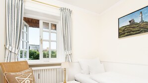 A cozy bedroom with a white bed, a wicker chair, a small bedside table with a lamp, and a window with curtains. - Rasmus 1 from A&M - Rasmus 1 (Kampen (Sylt))