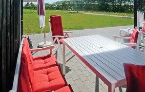 Un patio al aire libre con una mesa blanca y sillas rojas, una bandera y una zona verde de césped al fondo.