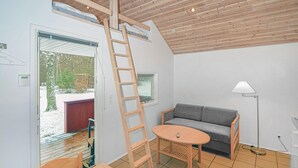 Ein Zimmer mit Holzboden, einem grauen Sofa, einem runden Holztisch, einer Leiter und einem Fenster mit Jalousien.