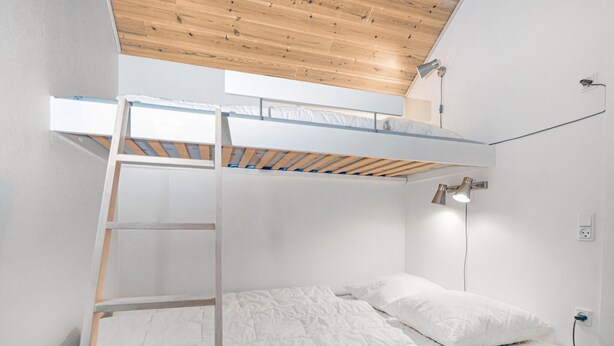 Ein Stockbett mit Holzlade, ein Einzelbett darunter und ein an der Wand befestigter Schreibtisch mit Lampe.