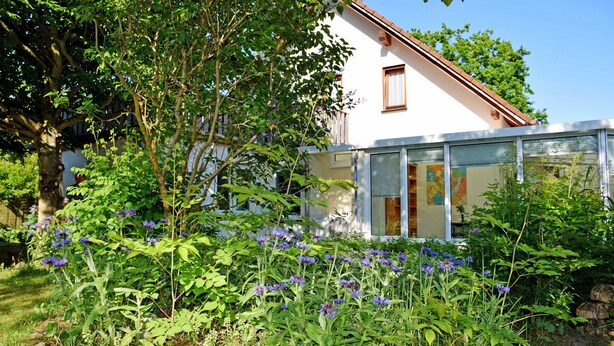 A modern house with a glass facade, surrounded by a garden with various plants and flowers. - Apartment 02 - Guesthouse Captain Winston (Putbus)