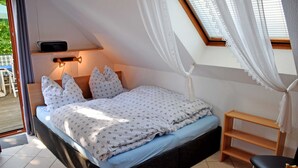 A bedroom with a bed, a bedside table, a shelf, and a window with curtains. - Apartment 02 - Guesthouse Captain Winston (Putbus)