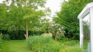 A lush garden with a variety of plants and trees, a well-maintained lawn, and a white building with large windows. - Apartment 02 - Guesthouse Captain Winston (Putbus)