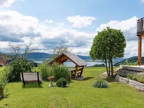 A wooden cabin with a sloped roof, a bench, and a tree in a grassy area with a view of mountains and a lake.