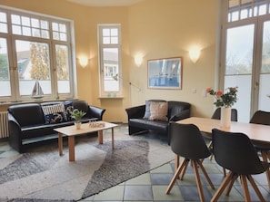 A living room with a sofa, a coffee table, a dining table with chairs, and a window with a sailboat model.