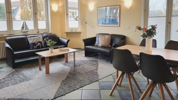 Un salon comprenant un canapé, une table basse, une table à manger avec des chaises et une fenêtre ornée d’une maquette de voilier.