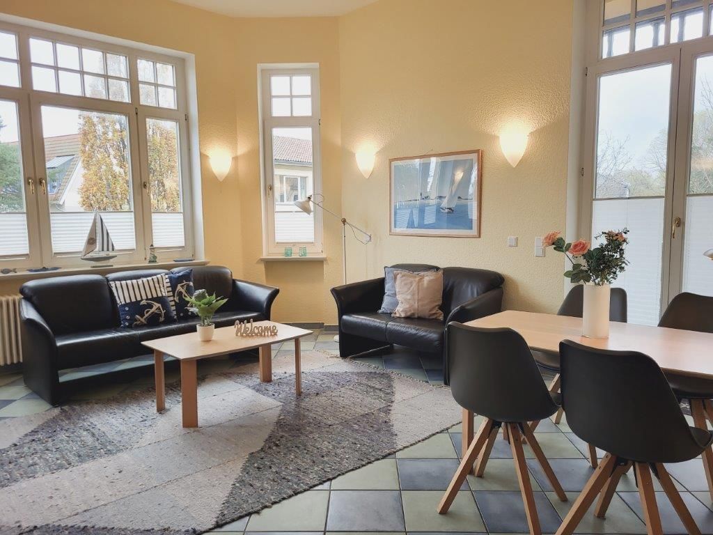 A living room with a sofa, a coffee table, a dining table with chairs, and a window with a sailboat model.