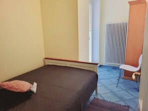 Una habitación pequeña con cama individual, pared sencilla, un radiador y una puerta.