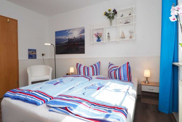 A hotel room with a bed, a chair, a desk with a lamp, a shelf with decorative items, and a painting on the wall.