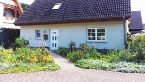 A house with a tiled roof, a front door, and a small window. There is a garden with various plants and flowers in front of the house.