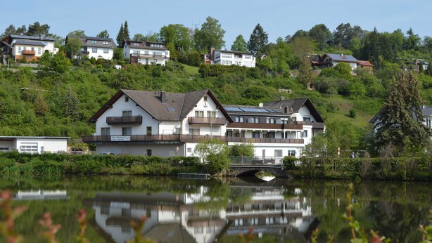 Une propriété au bord du lac, avec un pont, des bâtiments qui se reflètent dans l’eau et une végétation luxuriante.