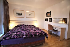 A bedroom with a bed, a desk, and a chair. There are framed pictures on the wall.