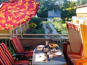 A balcony with a table set for breakfast, a red umbrella, and a view of a house and garden. - Zempin Ostseepark WE 30 Insel Usedom only 150m to the beach - WE 30 Strandläufer (Zempin)