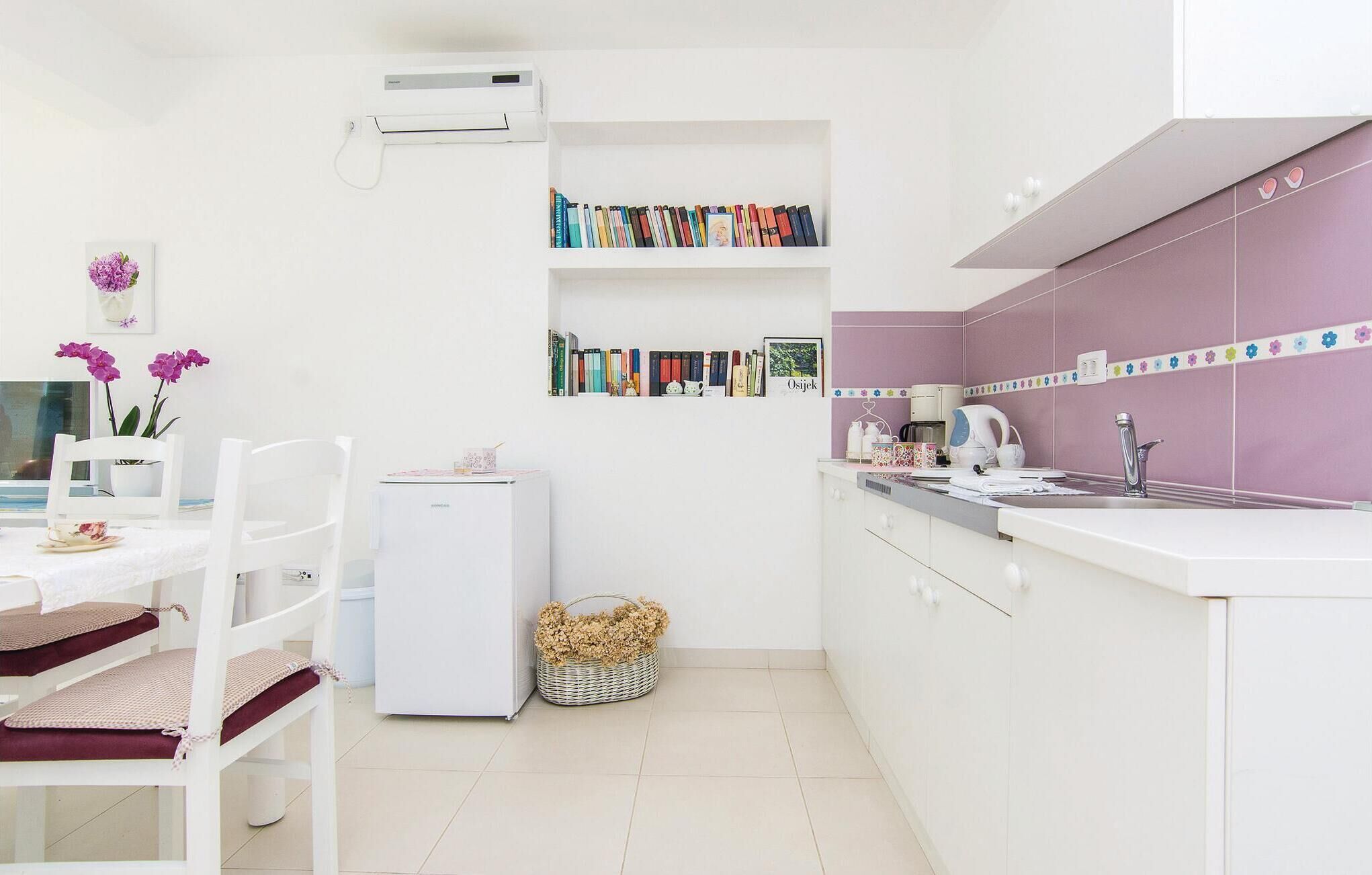 Een moderne keuken met witte kasten, een paars aanrechtpaneel en een witte koelkast. Er is een eethoek met een witte tafel en stoelen, en een klein wit schap met boeken en een ingelijste foto.