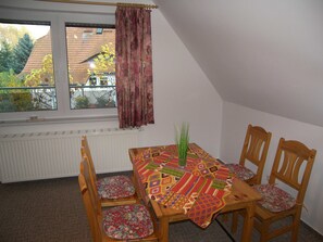 Ein Essbereich mit einem Tisch für vier Personen, Holzstühle mit floralen Kissen, ein Fenster mit Blick auf ein Haus und Bäume sowie ein Heizkörper unterhalb des Fensters.