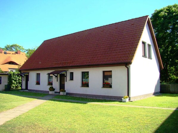 Ein eingeschossiges Haus mit rotem Ziegeldach, weißen Außenwänden und einer kleinen Veranda. Auf der Veranda stehen Kübelpflanzen und ein Weg führt zum Eingang.
