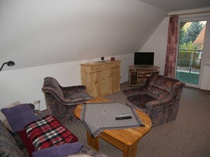 Ein Wohnzimmer mit zwei gemusterten Sesseln, einem Holztisch, einem Fernseher auf einem Schrank und einer Holzkommode.