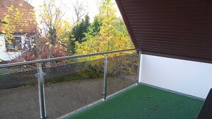 Un balcón con baranda de metal, césped artificial verde y vistas a árboles con hojas amarillas.