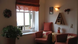 Una habitación acogedora con dos sillones, una mesita, una planta en maceta, una ventana con cortina, un estante en la pared y una lámpara de pared.