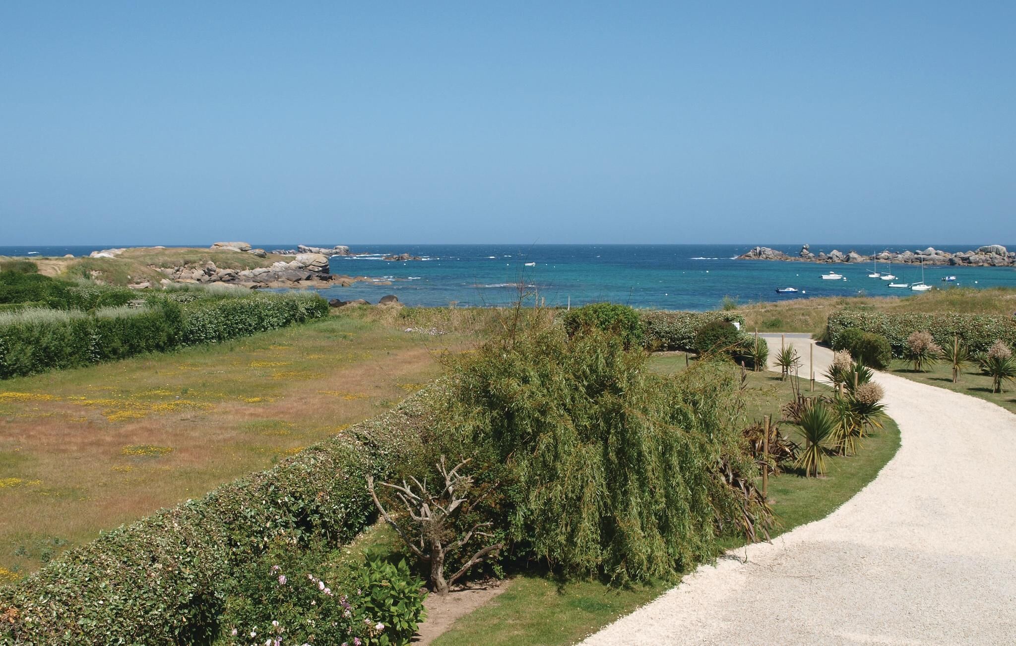 Un paisaje costero con un sendero de grava que conduce a una zona cubierta de hierba, con abundante matorral y vistas al mar con barcos.