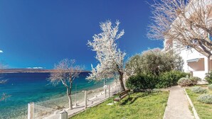 Vista costera con un mar azul cristalino, un edificio blanco con balcón y una zona verde con árboles.