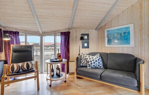 A cozy room with a wooden ceiling, a grey sofa, a wooden chair, a small round table with a bottle and glass, and a window with purple curtains. - Beautiful home in Hvide Sande with sauna (Hvide Sande)