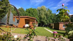 A rustic wooden cabin with a porch, surrounded by a well-maintained lawn and flowering plants.
