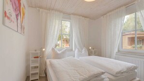 A bedroom with two beds, white curtains, a bedside table with a lamp, a shelf with books, and a window with a view of the outside.