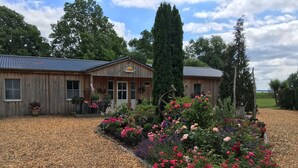 A wooden building with a sloped roof, surrounded by a garden with various flowering plants and a tall evergreen tree.