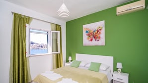 Ein Schlafzimmer mit Bett, grünen Vorhängen, einem Fenster mit Blick, einem farbenfrohen abstrakten Gemälde und einer weißen Klimaanlage.