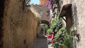 A narrow stone alleyway with a rustic stone wall on one side and a building with a wooden balcony on the other. - Bungalow in a holiday village (Castiglione della Pescaia)