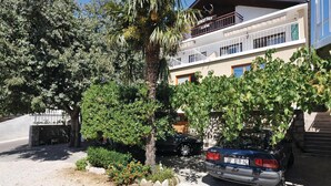 A residential building with a balcony, a parked car, and a garden area with various plants and a palm tree. - Nice apartment in Starigrad Paklenica (Starigrad Paklenica)