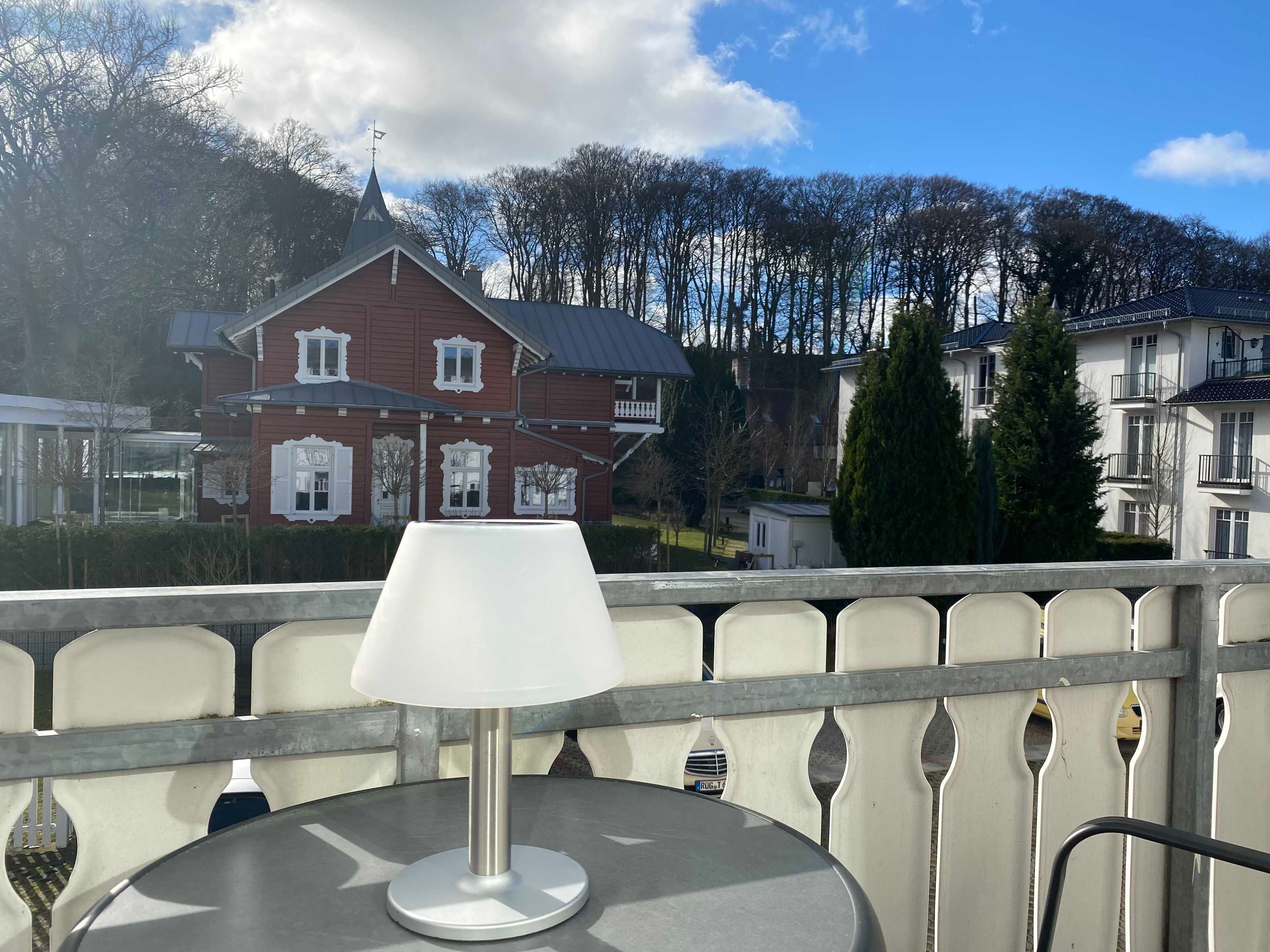 A balcony with a table and a lamp, overlooking a residential area with houses and trees.