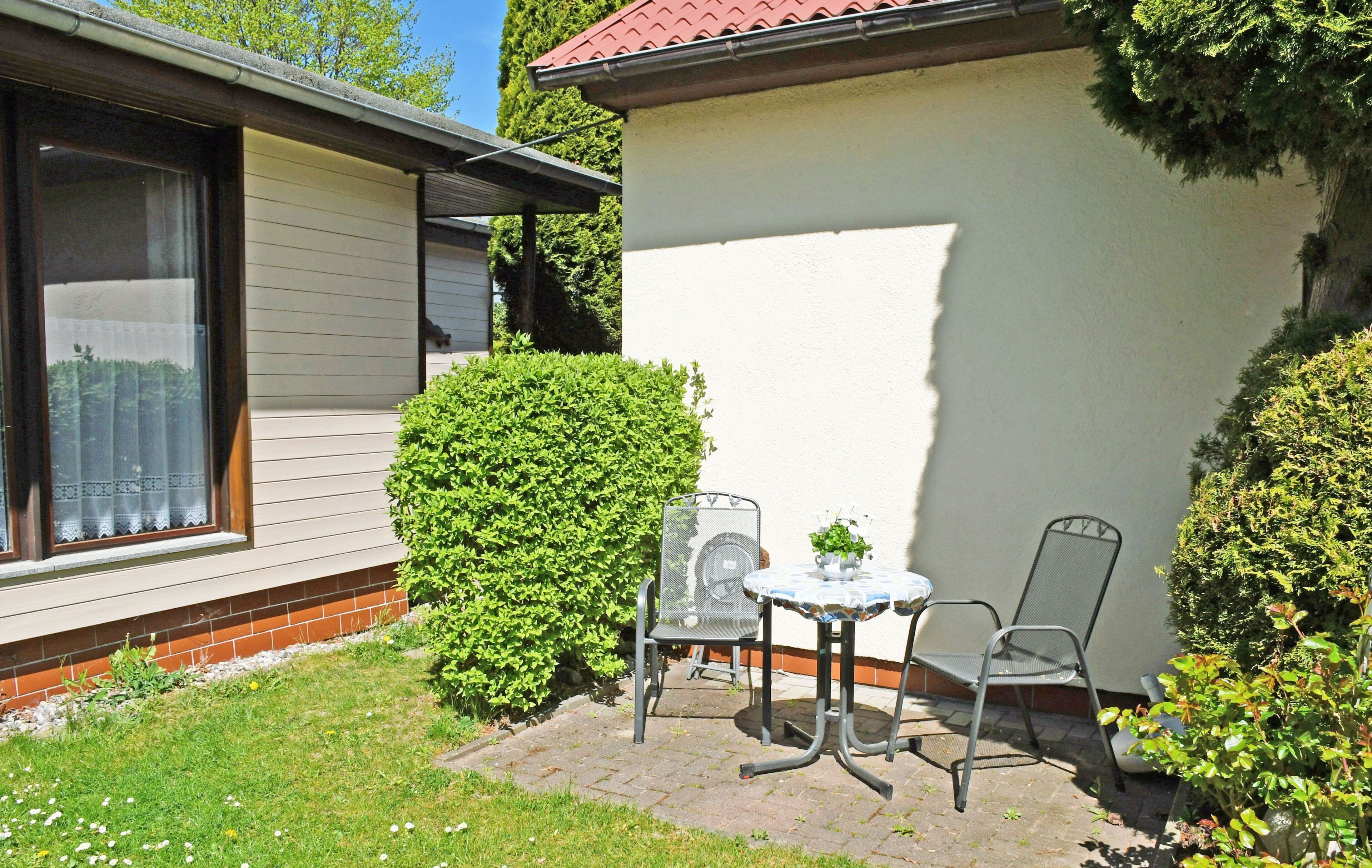 Une terrasse avec une table et des chaises, un petit arbuste et un bâtiment avec une fenêtre.