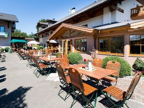 Un área de comedor al aire libre con mesas y sillas de madera, un espacio de estar techado y un edificio con ventanas y balcones amplios.